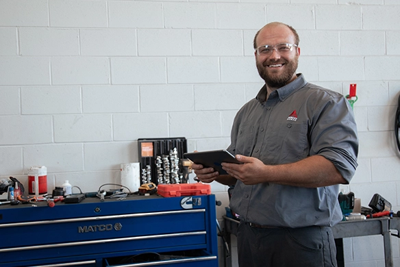 Smiling AGCO service technician holding a tablet in a workshop, standing next to a blue Matco tool chest.