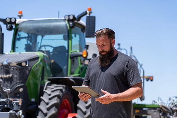 Farmer looking at iPad reading the latest news from AgRevolution, next to a Fendt tractor under a bright blue sky.